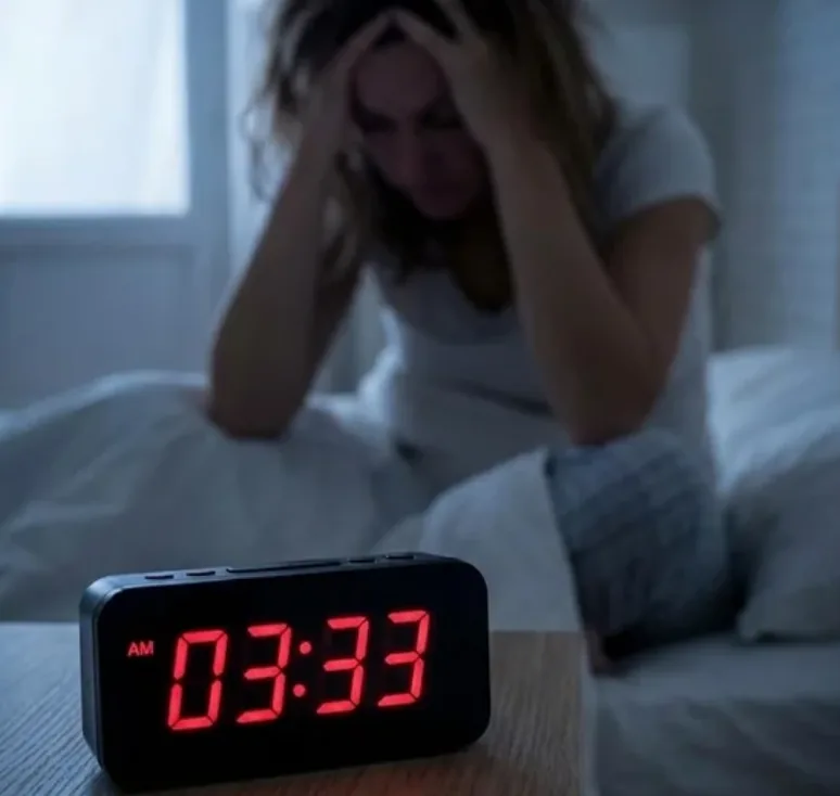 A digital clock reading 03:33 in the foreground while a woman sits awake in bed behind it